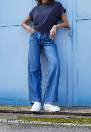 Blauwe wijde spijkerbroek, kort donkerblauw shirt en witte sneakers. De denim heeft een vervaagde wassing; het shirt heeft opgerolde mouwen. Op de achtergrond is een blauwe muur te zien.