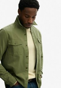 Grüne Baumwollbluse mit Stehkragen, zwei Brusttaschen und Knopfverschlüssen, getragen über einem hellen Pullover.