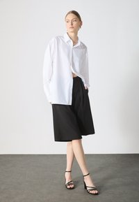 White button-up shirt, black tailored shorts, and strappy black heels. Smooth fabrics with a relaxed fit, featuring a pocket on the shirt.