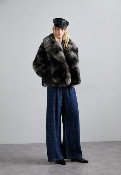 Γυναίκα που φοράει μαύρο δερμάτινο καπέλο, σκούφο πολύχρωμο ψεύτικο γούνα, φαρδιά μπλε τζιν παντελόνια και μαύρες μυτερές παπούτσια, standing σε γκρι δάπεδο.