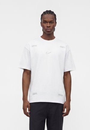 Junger Mann mit geflochtenem Haar, der ein schlichtes weißes Nike-T-Shirt und schwarze Hose trägt, steht vor einem weißen Hintergrund.