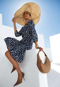 Navy-blaues Polka-Dot-Kleid mit Dreiviertelärmel, kombiniert mit einem großen Strohhut und braunen Absatzschuhen. Die braune Handtasche hebt den Look hervor.