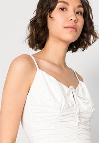 Top sem mangas branco com tecido franzido e detalhe de laço no peito, feito de material suave, apresentando alças finas e um corte descontraído.