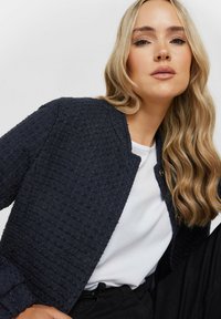 Giacca blu marina con texture, scollatura rotonda e due bottoni frontali, indossata su una camicia bianca, abbinata a pantaloni neri. Capelli lunghi e mossi.