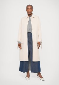 Vrouw in een lange beige jas over een grijze gebutton-up trui en een blauwe denim rok met witte hakken, staande tegen een witte achtergrond.