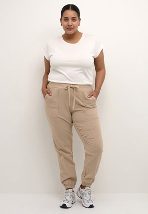 Beige joggingbroeken met elastische boorden en een trekkoord in de taille, gecombineerd met een fit wit T-shirt met korte mouwen. De textuur is zacht en casual.