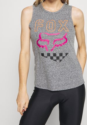 Kobieta w szarym topie bez rękawów z pomarańczowym napisem „FOX”, różowym logo głowy lisa i czarnym wzorem w kratę, ubrana w czarne legginsy.