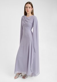 Lange lavendelkleurige jurk met gedrapeerde halslijn en vloeiende mouwen, gemaakt van lichtgewicht stof. Zilveren sandalen met banden maken de look af.