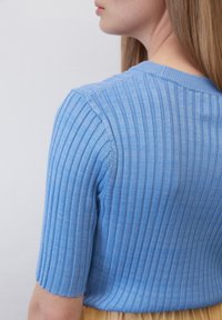 Světlomodrý pletený top s krátkým rukávem, který má kulatý výstřih a texturovaný povrch. Materiál vypadá měkký a lehce průhledný.