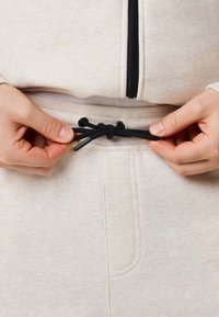 Ljusbeige hoodie med svart dragkedja, kombinerad med matchande sweatpants. Händer justerar den svarta dragskon i midjan. Mjuk textur.