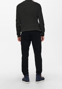 Dunkelgrüner, gerippter Pullover, schwarze Hose und marineblaue Sneakers. Der Pullover hat einen Rundhalsausschnitt und lange Ärmel; die Hose hat eine lockere Passform.