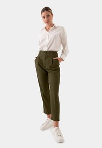 Olive green high-waisted trousers with side pockets and a front button closure, paired with a white striped button-up shirt and white sneakers.