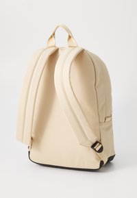 Rucsac bej cu o textură netedă, design curbat, bretele ajustabile și un accent subtil al logo-ului. Dispune de un mâner superior și o pocket laterală.