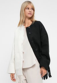 Mulher com cabelo loiro vestindo um casaco cardigã preto e branco abotoado com franjas, por cima de calças claras e uma blusa branca, olhando para o lado.