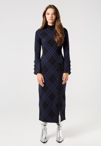 Lungo vestito in maglia nera con motivo a quadri blu scuro; presenta maniche lunghe, bottoni laterali e uno spacco frontale. Indossato con stivali alti fino al ginocchio di colore argento lucido.
