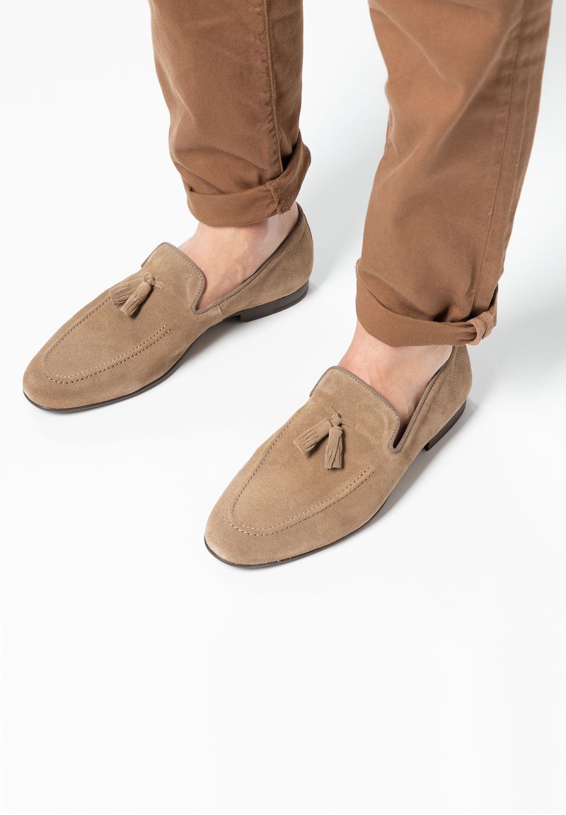 Suède loafers in lichtbruin, met een franjesaccent, ronde neus en gestikte details. Gecombineerd met opgerolde bruine broeken.