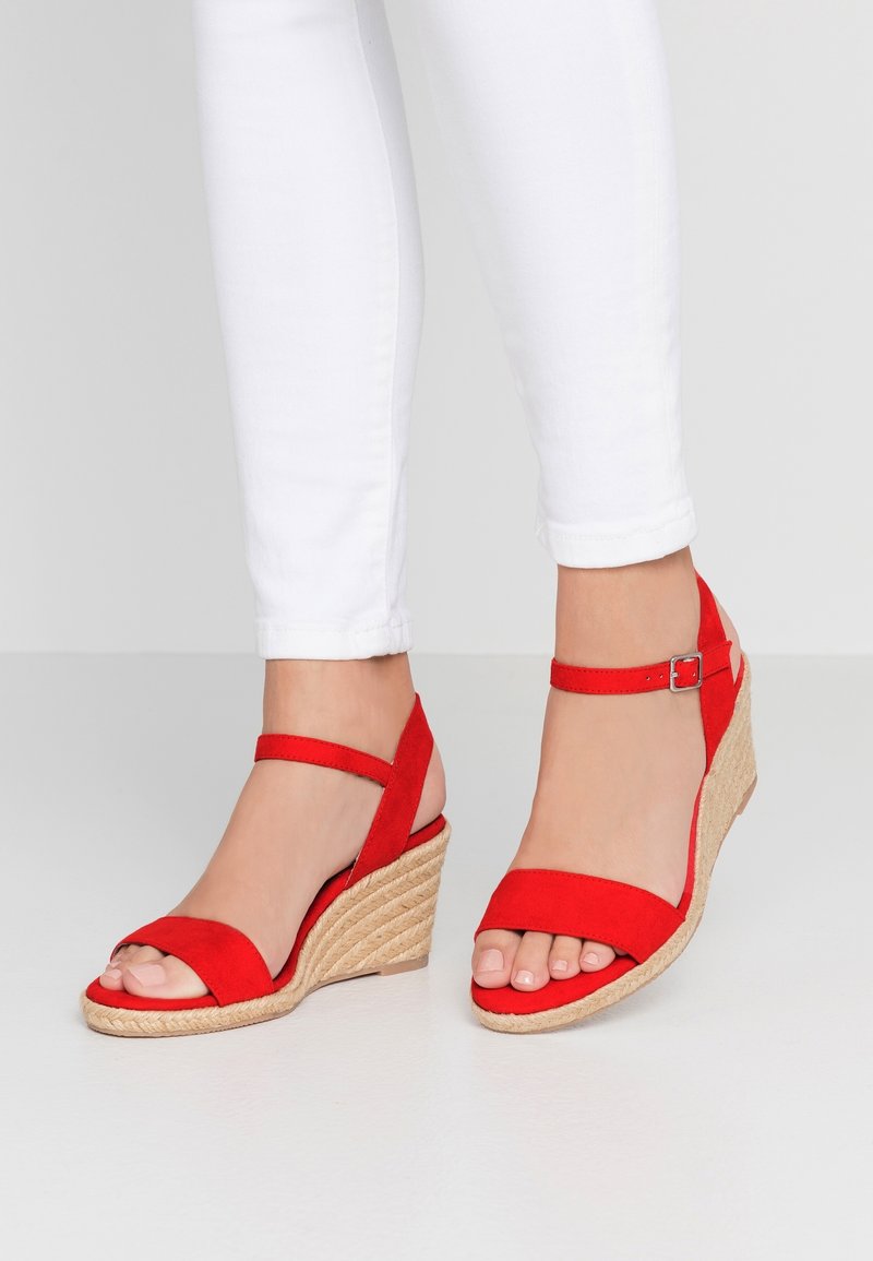 Pieds portant des sandales compensées rouges avec des brides à la cheville et un pantalon blanc ajusté sur un fond clair uni.