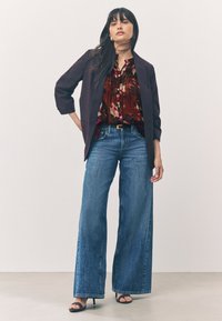 Blazer viola scuro, camicetta floreale rossa e jeans blu a gamba larga. L'outfit presenta una texture liscia, bottoni sulla camicetta e una cintura.