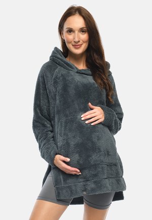 Schwangere Frau trägt übergroßen dunkelgrauen Fleece-Hoodie und graue Shorts, hält sanft ihren Bauch und lächelt.