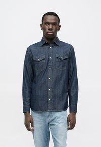 Tamsiai mėlyna džinsinė palaidinė su ilgomis rankovėmis, dviem krūtinės kišenėmis ir baltomis sagomis. Turi klasikinio stiliaus apykaklę ir laisvo stiliaus prigludimą.