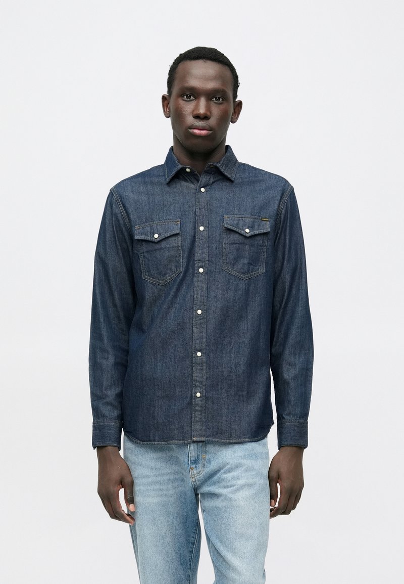 Tamsiai mėlyna džinsinė palaidinė su ilgomis rankovėmis, dviem krūtinės kišenėmis ir baltomis sagomis. Turi klasikinio stiliaus apykaklę ir laisvo stiliaus prigludimą.