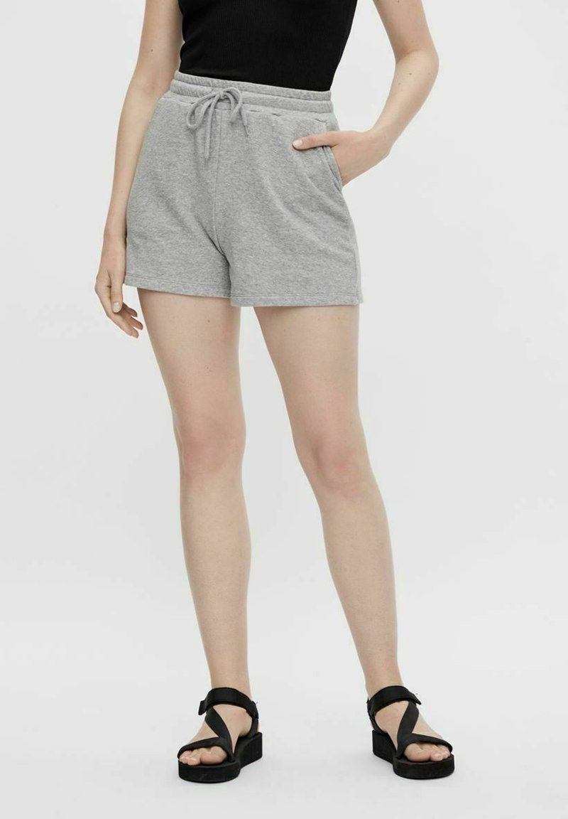 Shorts en coton gris avec une ceinture élastique et un cordon de serrage, dotés de poches latérales. Portés avec des sandales plates noires, modèle debout sur un fond uni.