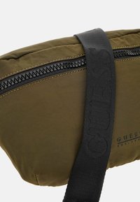 Khakifarbenes Nylon-Tasche mit einem strukturierten schwarzen Gurt, ausgestattet mit einem schwarzen Reißverschluss und einem geprägten Logo. Leicht gepolstert und mit einer geschwungenen Form.