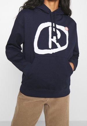 Persona che indossa una felpa con cappuccio blu navy con un grande logo astratto bianco e le mani nella tasca anteriore, abbinata a pantaloni di velluto a coste marrone chiaro.