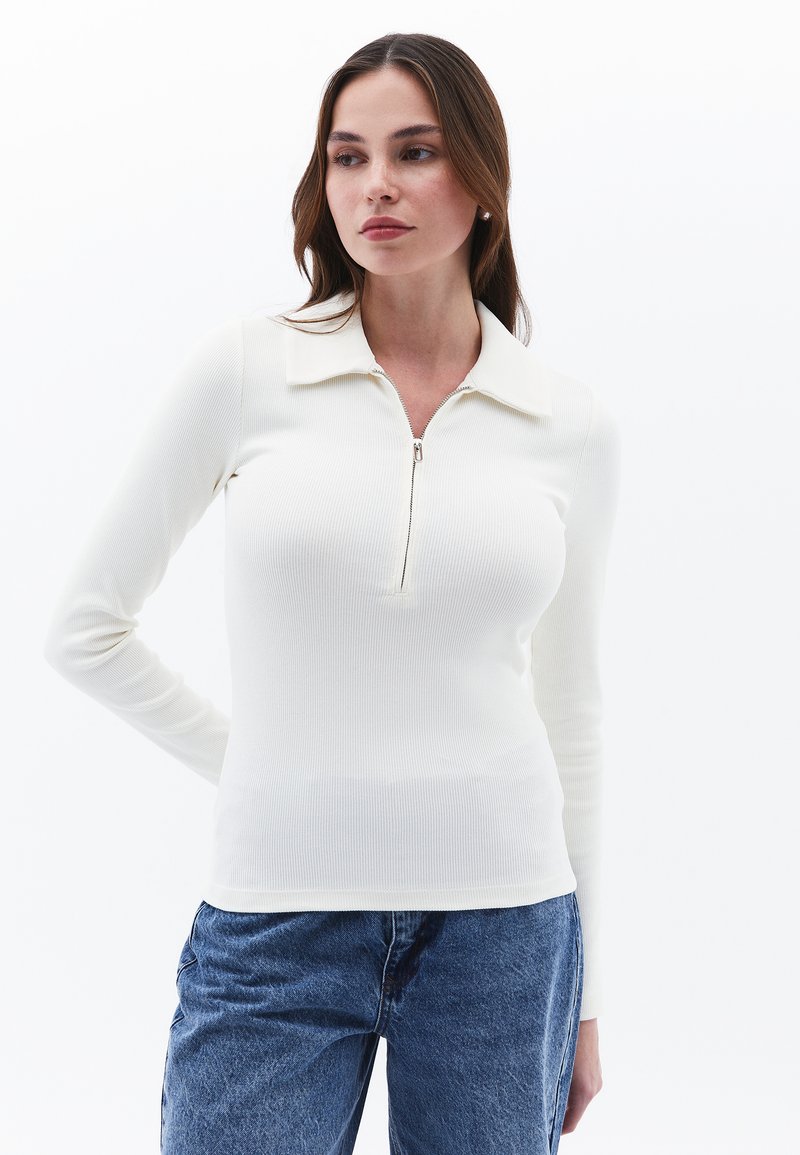 Biała, prążkowana bluzka z długim rękawem i zamkiem przy kołnierzu. Miękka tekstura i dopasowany krój, w połączeniu z dżinsami, tworząca swobodny wygląd.