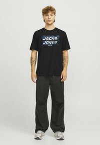 Sort T-shirt med "JACK & JONES" grafik i blå, båret med løse mørke bukser og grå sneakers. Modellen har lysebrunt hår og tatoveringer.