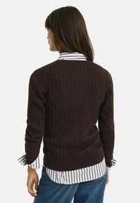 Dunkelbrauner gestrickter Pullover mit einem Zopfmuster, ausgestattet mit gerippten Bündchen. Über einem weißen Hemd mit dunklen Streifen und blauen Jeans getragen.