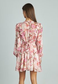 Vestido floral con un color rosa claro y acentos con volantes, mangas largas, un cinturón ajustable en la cintura y un diseño fluido hasta la rodilla con un patrón detallado.