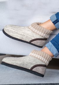 Wollen pantoffels met een beige textuur aan de bovenkant, geribbelde beige boord, bruine accenten en een zwarte rubberen zool; ontworpen voor comfort en warmte.