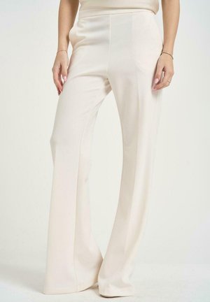 Donna che indossa pantaloni color crema a vita alta e gamba larga con le mani rilassate lungo i fianchi su uno sfondo chiaro uniforme.