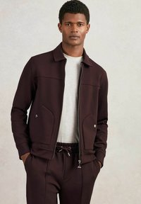 Geacă zip-up vișinie cu două buzunare în față și guler, asortată cu pantaloni vișinii și o cămașă gri deschis dedesubt.