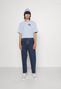 Lichtblauw oversized t-shirt met golfprint, gecombineerd met losse marineblauwe jeans en witte hoge sneakers. Model draagt een lila beanie.