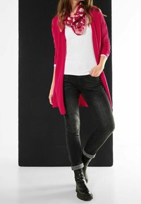 Lång rosa cardigan över en vit skjorta, kombinerad med mörka jeans och svarta ankelstövlar. En mönstrad rosa scarf tillför detaljer. Släta tygtexturer.