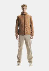 Joven de pie, vistiendo una chaqueta con capucha marrón, pantalones cargo beige y zapatillas blancas, mirando hacia adelante contra un fondo sencillo.