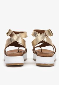 Złote metaliczne sandały z krzyżującymi się paskami, brązową amortyzowaną wkładką oraz białą masywną podeszwą. Zapięcie na klamrę na pasku wokół kostki.