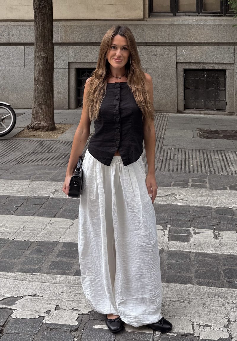 Mujer con el pelo largo, vestida con un top negro sin mangas, pantalones blancos anchos y zapatos negros, de pie en una acera de la ciudad sosteniendo una pequeña bolsa negra.