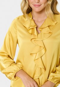 Gelbe Satinbluse mit rüschentFront, langen Ärmeln mit elastischen Bündchen und V-Ausschnitt. Glatte Textur und lebendige Farbe.