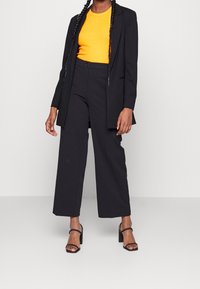 Costume noir avec un long blazer, un haut orange et un pantalon à jambes larges. Présente une texture lisse et des sandales noires à talons hauts.