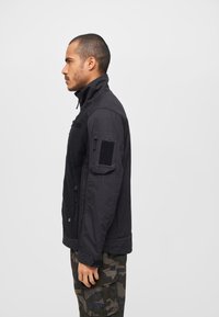 Black jacket with a high collar, zippered front, arm pocket with Velcro, and textured fabric; worn with camouflage cargo pants.