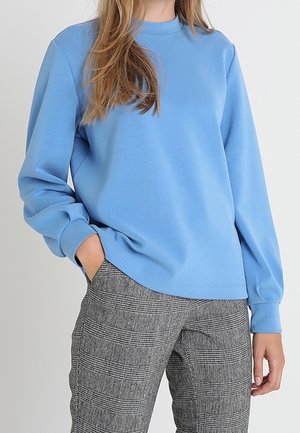 Osoba ubrana w jasnoniebieską bluzę z długim rękawem i szare spodnie w kratę, z jedną ręką w kieszeni, na neutralnym tle.