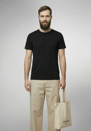Homme barbu portant un t-shirt noir et un pantalon beige tient un sac fourre-tout beige avec le texte "ERVERTE Paris" sur un fond uni.
