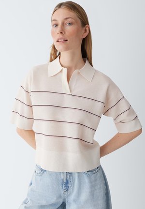 Junge Frau mit langen blonden Haaren, die ein cremefarbenes Poloshirt mit dünnen horizontalen Streifen und hellblaue Jeans trägt, posiert mit den Händen in der Hüfte.