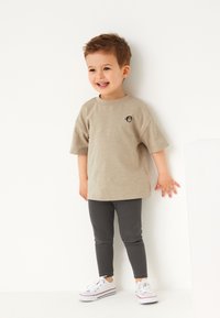 Beiges T-Shirt mit kurzen Ärmeln und schwarzem Affenlogo, kombiniert mit grauen Leggings und weißen Turnschuhen. Kind steht vor einer weißen Wand.