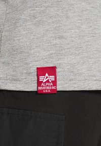 Tessuto grigio con un'etichetta rossa che mostra "ALPHA INDUSTRIES INC. U.S.A." L'etichetta presenta un logo con una A astratta e tre linee orizzontali.