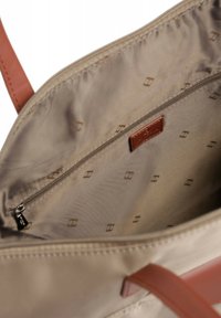 Interno di una borsa beige con una texture liscia, dotato di una tasca con zip e di una toppa con logo marrone. Foderato con un tessuto stampato.