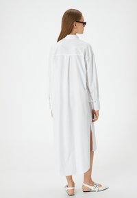 Weißes langes Hemdkleid mit Rückenpasse, seitlichen Schlitzern und geknöpften Manschetten, aus leichtem Stoff gefertigt. Glatte Textur, minimalistisches Design.